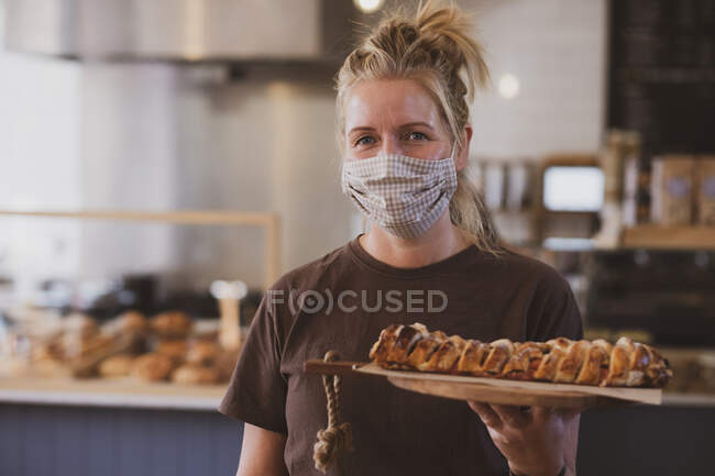 Cameriera bionda che indossa maschera viso lavorando in un caffè, portando piatto di cibo. — Foto stock
