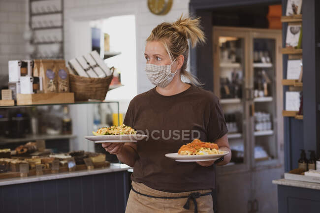 Blonde Kellnerin mit Mundschutz arbeitet in einem Café und trägt Teller mit Lebensmitteln. — Stockfoto