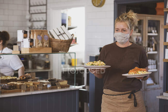 Blond waitress wearing face mask working in a cafe, carrying plates of food. — Stock Photo