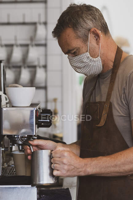 Barista homme portant tablier marron et masque facial travaillant dans un café, moussant lait. — Photo de stock