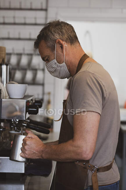 Varón barista con delantal marrón y mascarilla facial trabajando en un café, leche espumosa. - foto de stock