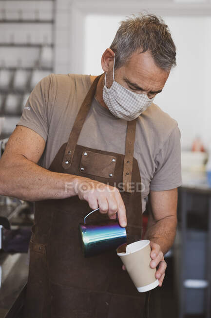 Männlicher Barista mit brauner Schürze und Gesichtsmaske arbeitet in einem Café und schenkt Kaffee ein. — Stockfoto