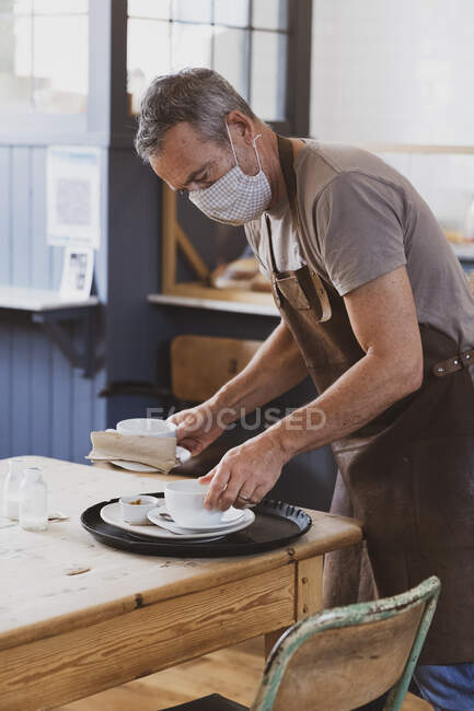 Офіціант у коричневому фартуху та масці для обличчя, що працює в кафе, чистий стіл . — стокове фото