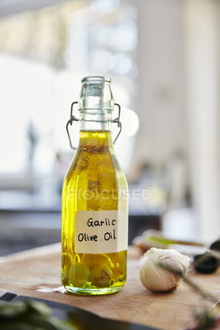 Стеклянная бутылка с оливковым маслом и чесноком, стоящая на доске для рубки на кухне — стоковое фото