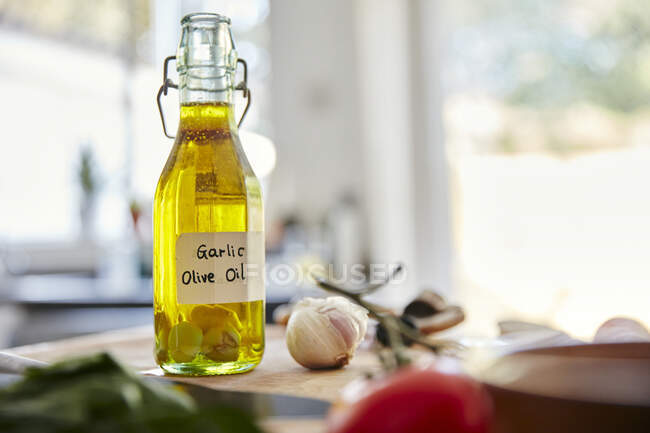 Glass bottle containing olive oil and garlic cloves standing on chopping board in kitchen — Stock Photo