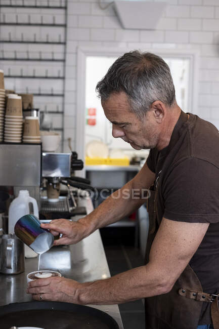 Männlicher Barista trägt braune Schürze, steht an der Espressomaschine und gießt Milch ein. — Stockfoto
