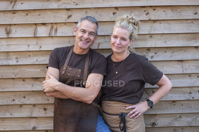 Waiter with short grey hair and waitress with blond hair, both wearing brown aprons, leaning against wooden wall, smiling at camera. — Stock Photo