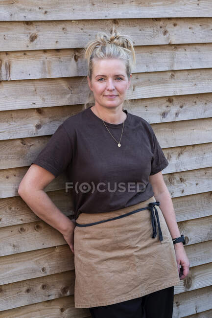 Portrait of waitress wearing brown apron, leaning against wall, smiling. — Stock Photo