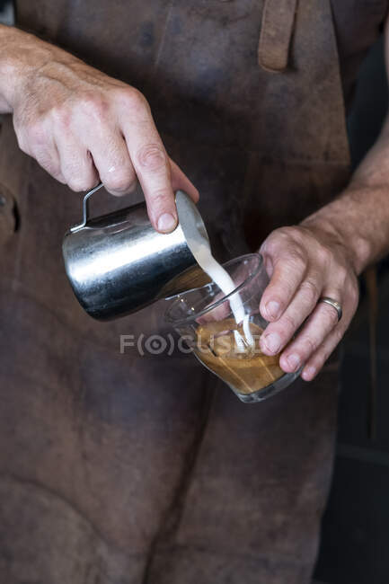 Primer plano de barista vistiendo delantal marrón vertiendo café con leche. - foto de stock