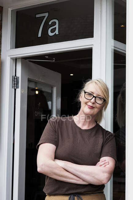 Portrait de serveuse aux cheveux blonds et lunettes, portant un tablier marron, appuyée contre la porte d'entrée, souriant à la caméra. — Photo de stock