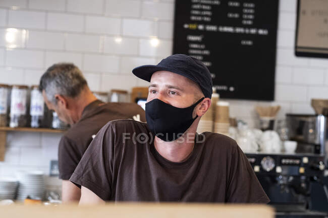Чоловіча бариста в чорній бейсбольній шапці та масці для обличчя, що працює за лічильником . — стокове фото