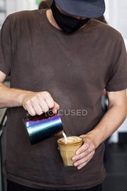 Close up of male barista wearing face mask pouring cafe latte. — Stock Photo