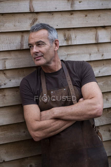 Retrato de barista masculino con pelo gris corto, con delantal marrón, brazos cruzados, apoyado contra pared de madera. - foto de stock