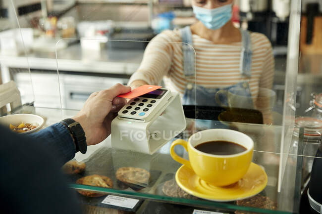 Жінка в масці за кафе з безпечним екраном, пропонуючи безконтактний платіжний термінал для клієнта. — стокове фото