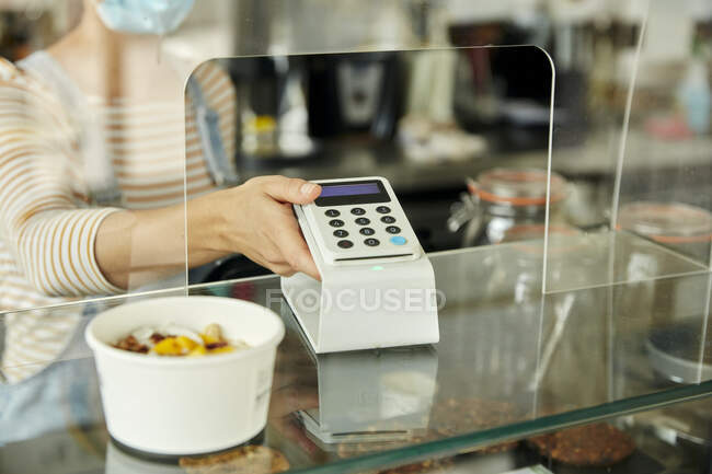 Woman in a face mask behind cafe counter with safety screen, offering a contactless payment terminal to a customer — Stock Photo