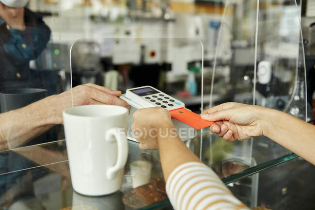 Женщина за стойкой кафе держит в руках бесконтактное платежное устройство, в то время как клиент использует мобильный телефон для оплаты счетов — стоковое фото