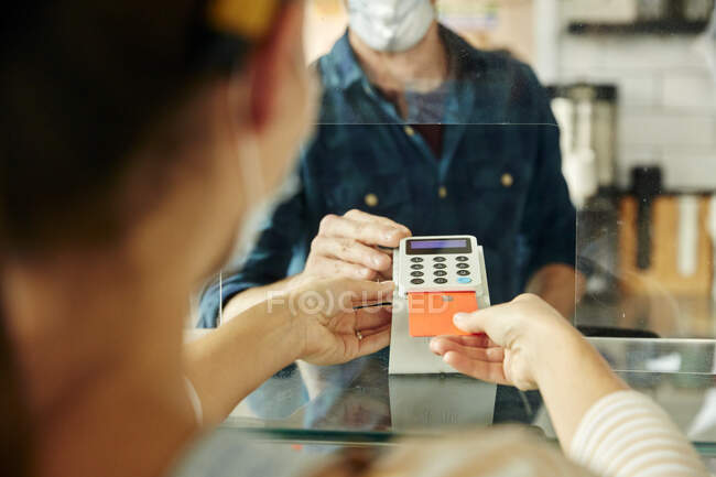 Woman wearing face mask behind cafe counter holding contactless payment device while customer uses mobile phone to pay bill — Stock Photo