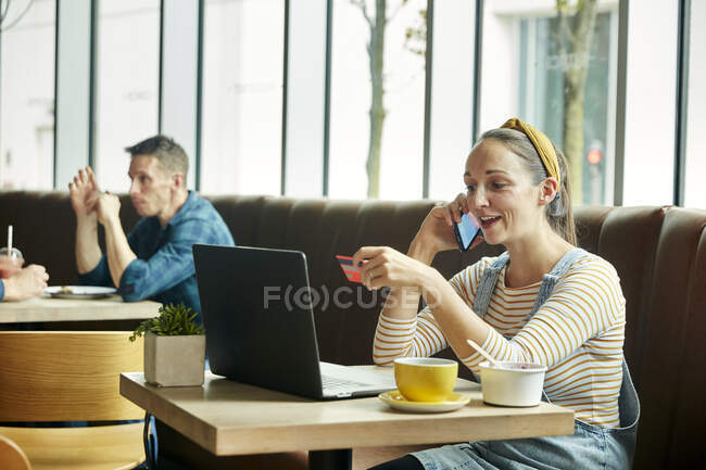 Frau sitzt mit Laptop in Café und telefoniert mit Smartphone — Stockfoto