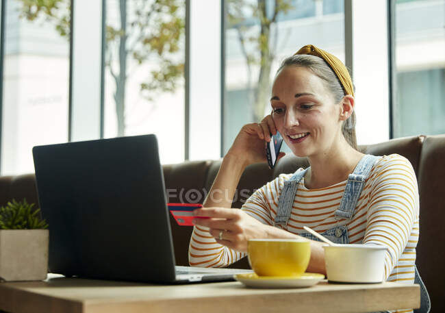 Mulher sentada em um café usando um laptop e falando em um telefone inteligente, segurando um cartão de crédito — Fotografia de Stock