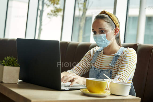 Mujer con una máscara facial sentada en un café usando una computadora portátil - foto de stock
