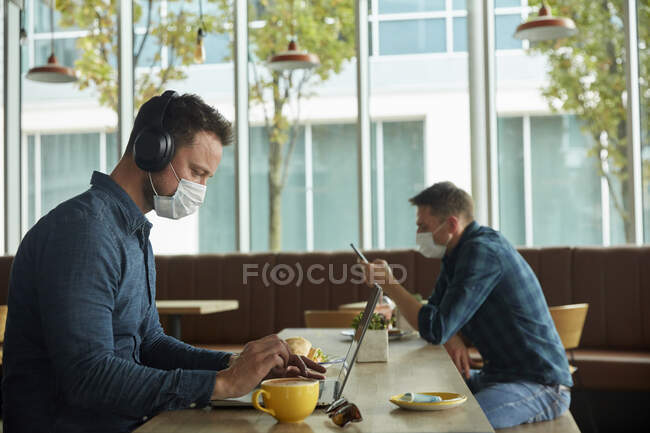 Dos hombres sentados en un café, trabajando remotamente con un portátil y un teléfono inteligente - foto de stock