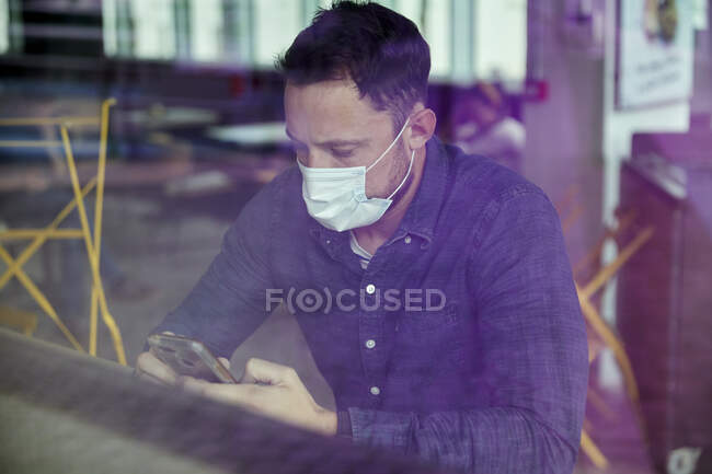 Hombre con una máscara facial sentado en una mesa de café usando un teléfono móvil, vista a través de una ventana - foto de stock
