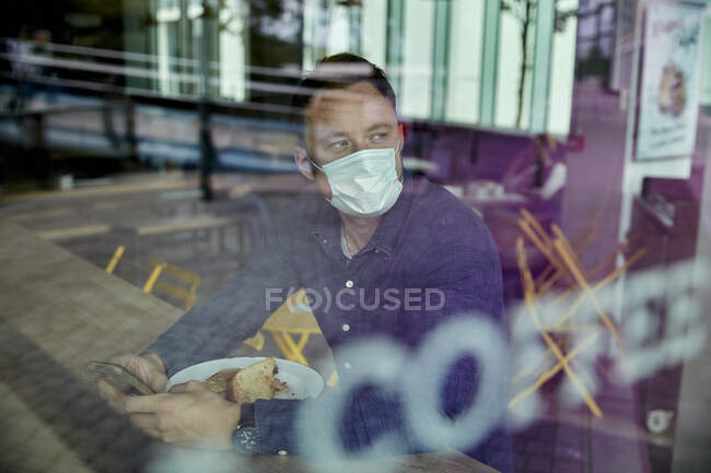Чоловік у масці для обличчя, що сидить за столом у кафе, перегляд через вікно — стокове фото