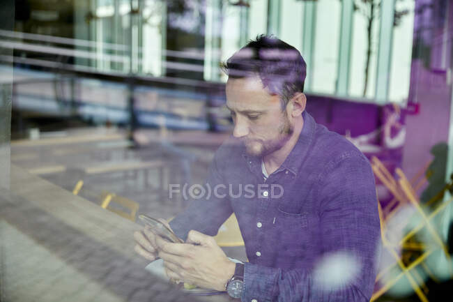 Homem em uma máscara facial sentado em uma mesa de café usando um telefone celular, vista através de uma janela — Fotografia de Stock