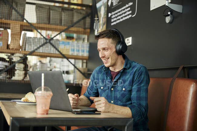 Чоловік сидить у кафе за допомогою ноутбука, носить навушники, приймаючи онлайн дзвінок . — стокове фото