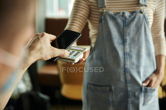 Женщина в маске для лица за стойкой кафе держит бесконтактное платежное устройство, в то время как клиент использует мобильный телефон для оплаты счетов — стоковое фото