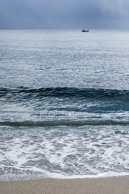 Meereslandschaft mit Sandstrand und Wellen unter stürmischem Himmel, Fischerboot in der Ferne. — Stockfoto