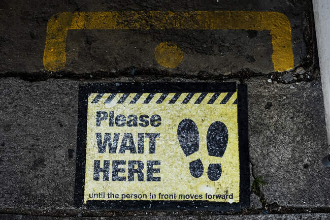 Alto angolo da vicino di nero e giallo 'Attendere qui' segno su asfalto terra. — Foto stock
