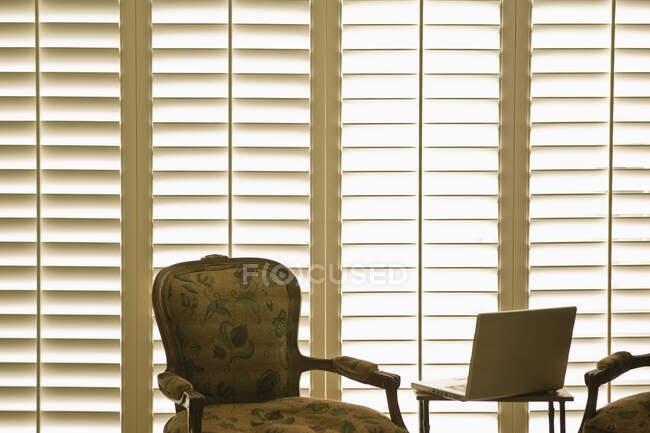 Armchair and laptop in front of domestic wooden window shutters. — Stock Photo