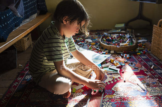 Garçon assis parmi les jouets sur le sol de sa chambre dans un patch de lumière du soleil — Photo de stock