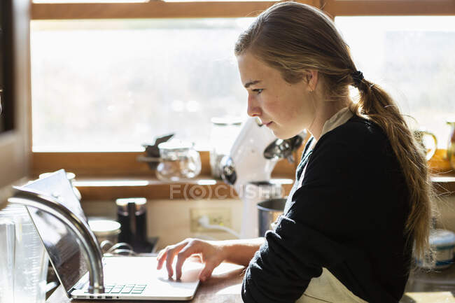 Teenage girl in a kitchen following a baking recipe on a laptop. — Stock Photo
