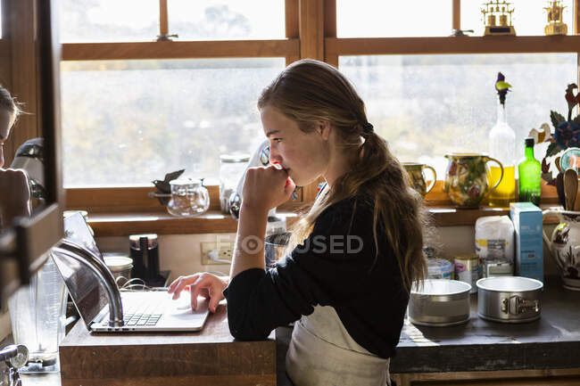 Adolescente dans une cuisine suivant une recette de cuisson sur un ordinateur portable. — Photo de stock