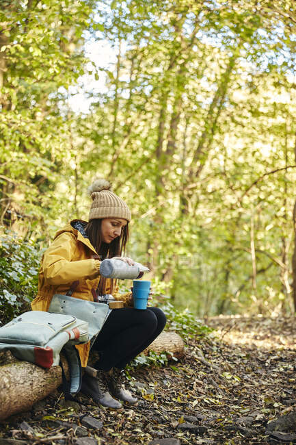 Женщина, сидящая на стволе дерева в лесу наливая напиток из термос фляжки — стоковое фото