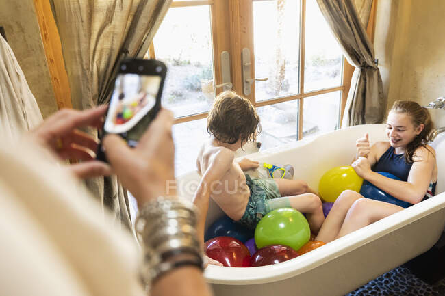 Persona che scatta una fotografia di smart phone di un ragazzo e sua sorella maggiore nella vasca da bagno con palloncini d'acqua — Foto stock