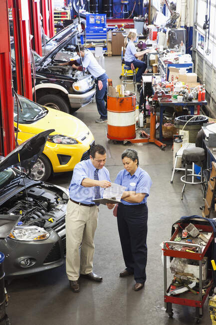 Proprietário da oficina de reparação de automóveis conversando com a mecânica feminina hispânica como colegas que trabalham no fundo — Fotografia de Stock