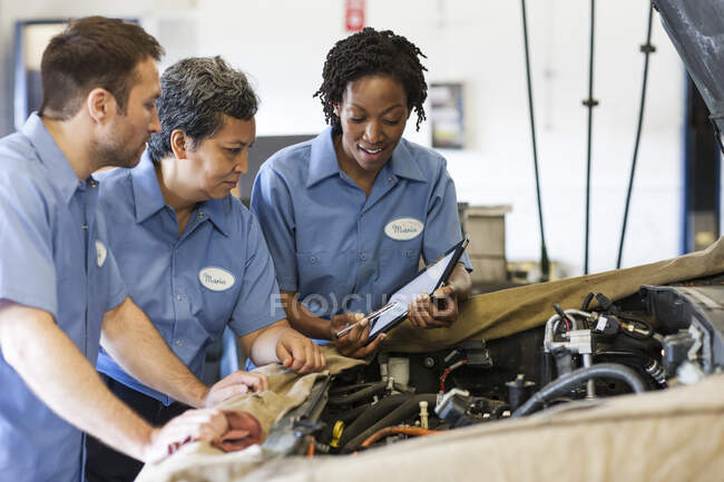 Three mechanics sharing a digital tablet and planning work on a car in for repair — Stock Photo