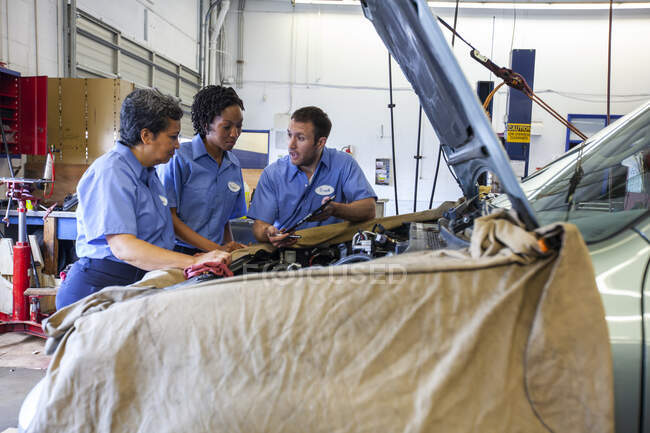Tres mecánicos compartiendo una tableta digital y planeando el trabajo en un coche para su reparación - foto de stock