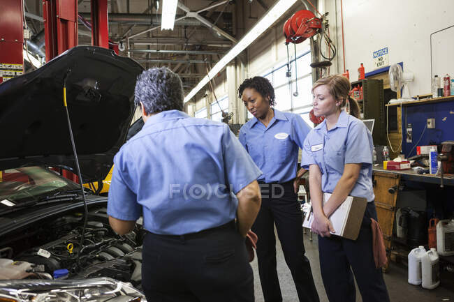 Trois mécaniciennes parlent en regardant à l'intérieur du compartiment moteur d'une voiture dans un atelier de réparation — Photo de stock