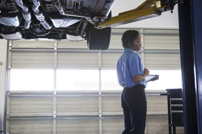 Mecánica femenina sosteniendo una tableta digital mientras está de pie debajo de un coche en un ascensor en un taller de reparación de automóviles - foto de stock