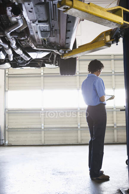 Female mechanic holding a digital tablet as she stands under a car on a lift in an auto repair shop — Stock Photo