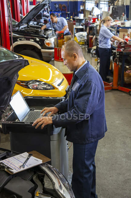 Pacific Islander mechanic typing on a laptop while co-workers work on cars in background — Stock Photo