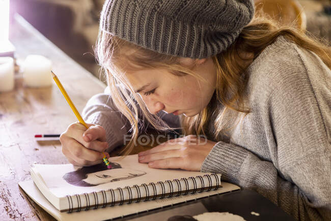 Teenage girl in a woolly hat drawing with a pencil on a sketchpad. — Stock Photo