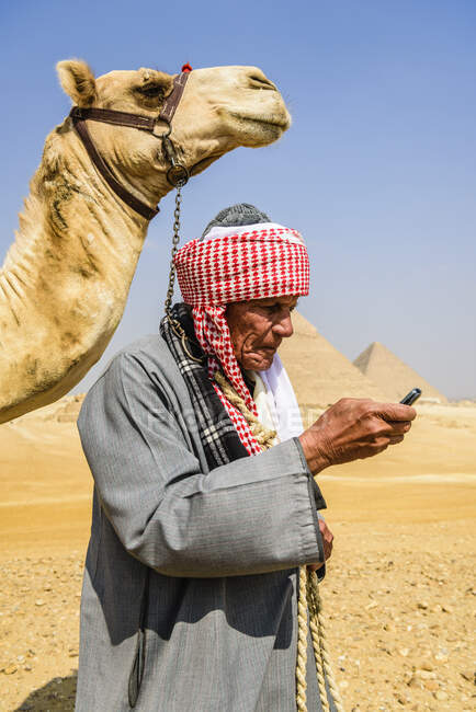Una guía con vestimenta tradicional árabe, bata y turbante usando un teléfono móvil, y su camello. - foto de stock
