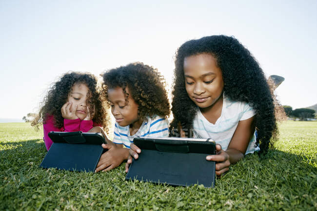 Chica joven de raza mixta y su hermano y hermana tumbados en la hierba con dos tabletas digitales, tiempo de pantalla - foto de stock