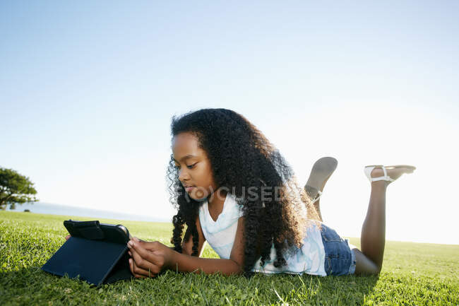 Neunjähriges Mischlingsmädchen liegt im Gras und blickt auf ein digitales Tablet. — Stockfoto