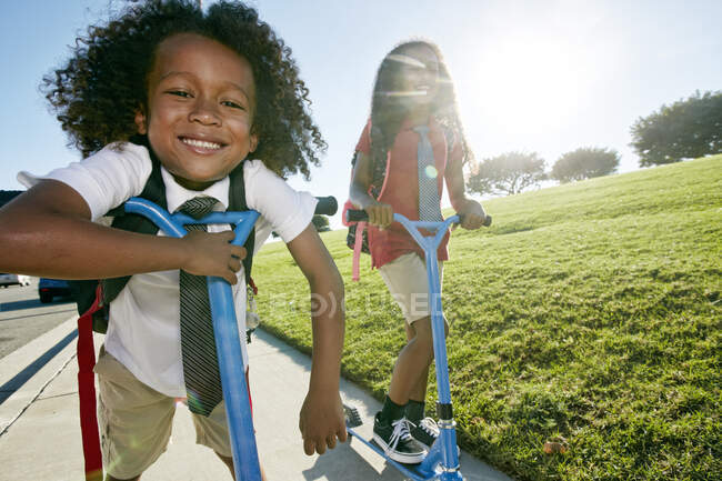 Симпатичный улыбчивый мальчик и его старшая сестра на скутерах по дорожке — стоковое фото